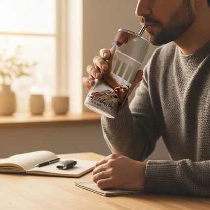 A man in a gray sweater is sitting at a desk, sipping from a straw-style beverage container with a printed design on it. There are various objects on the desk, including a notebook, a pen, and a cell phone.