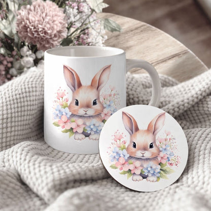 A white ceramic mug with a bunny illustration and a matching coaster featuring a bunny illustration are placed on a white blanket with pink flowers.