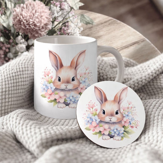 A white ceramic mug with a bunny illustration and a matching coaster featuring a bunny illustration are placed on a white blanket with pink flowers.