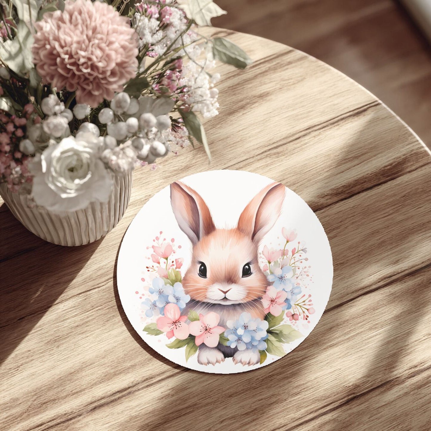 a round white plate with a picture of a bunny surrounded by flowers, placed on a wooden table.