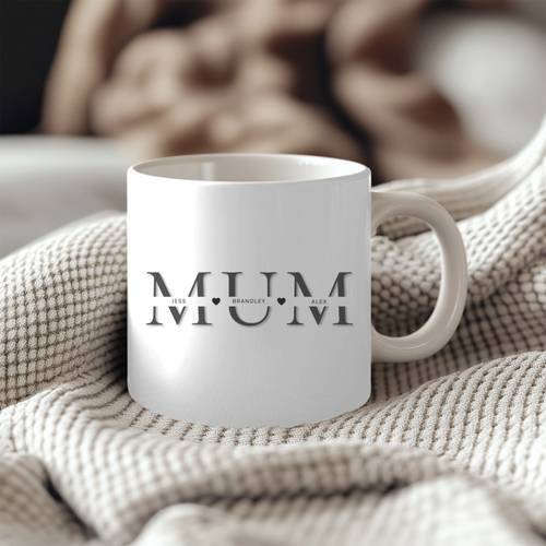 A white mug with the word "MUM" printed on it, sitting on a beige blanket.